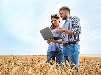 Frau und Mann stehen zusammen in einem Weizenfeld und der Mann hält einen Laptop in der Hand.