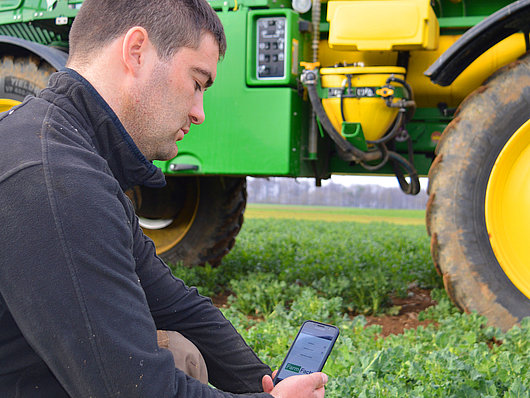 NEXT Farming Kunde Lohnunternehmer Christian Weik ist in der Hocke neben seinem Selbstfahrer zur Pflanzenschutz Behandlung und hält ein Smartphone in der Hand.