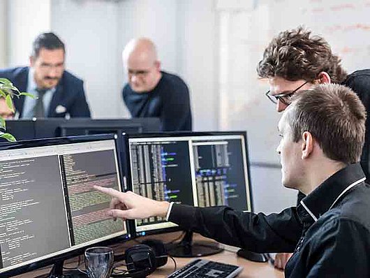 In einer Computerzentrale befinden sich zwei Männer vor zwei Bildschirmen im Vordergrund und ebenso zwei Männer vor zwei Bildschirmen im Hintergrund.