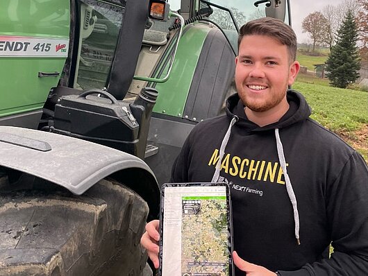 Junglandwirt Tobias Troppmann steht vor einem Fendt-Traktor und hält ein Tablet mit NEXT Farming Live in die Kamera