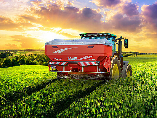Traktor mit Düngerstreuer bei Sonnenuntergang auf einem Weizenfeld.