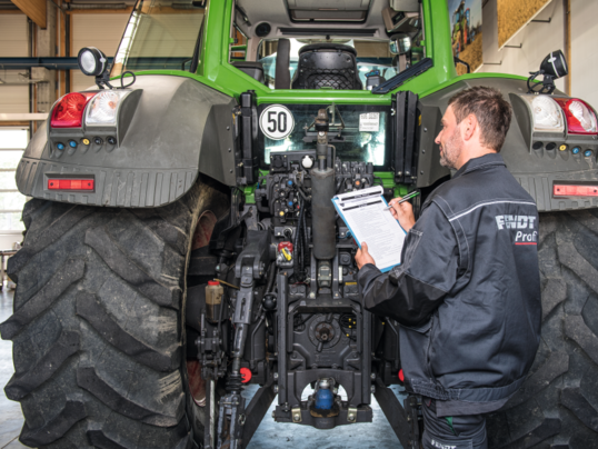 Ein Landtechnikhändler steht hinter einem Fendt-Traktor mit einem Klemmbrett und Stift in der Hand